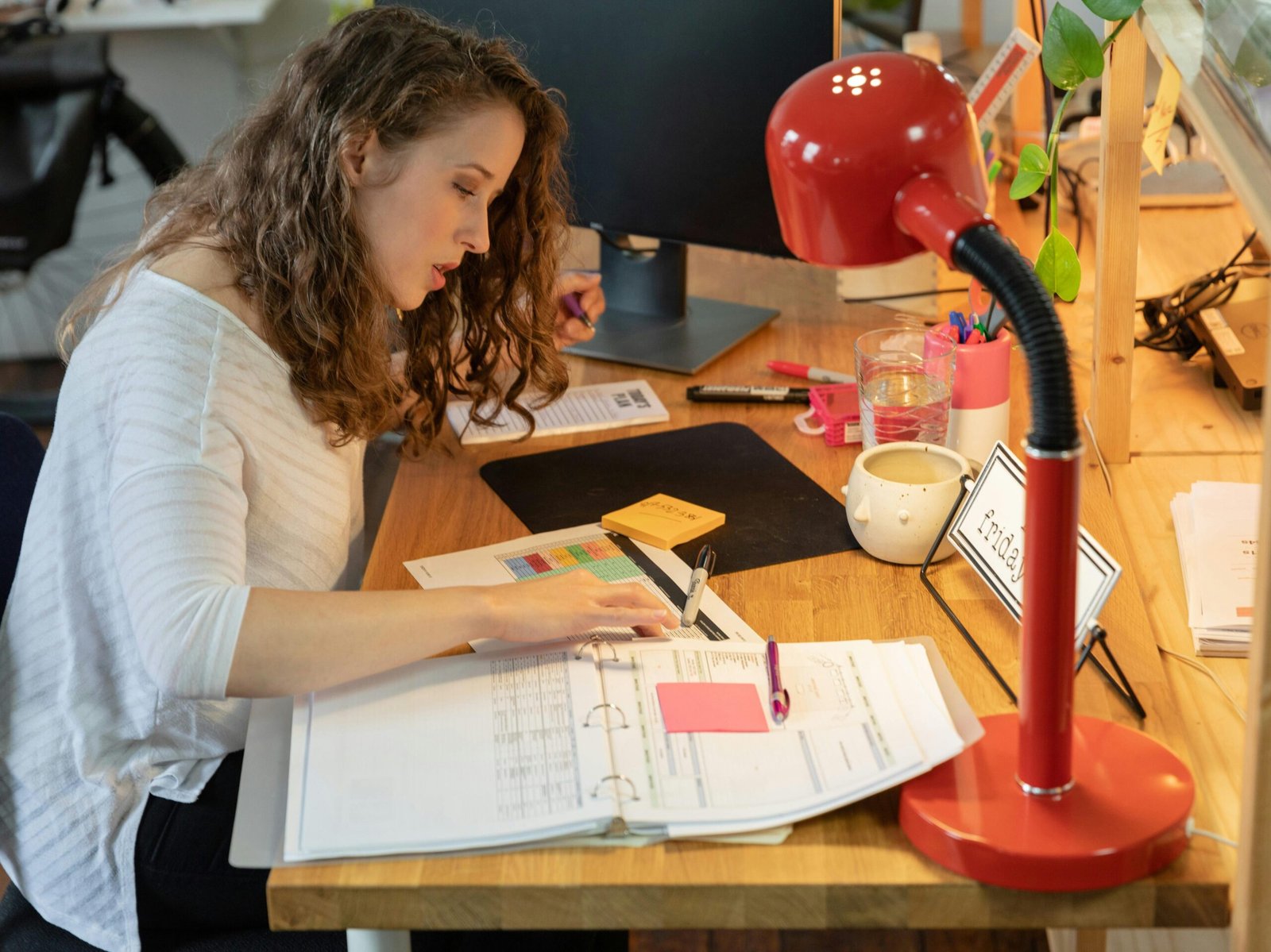 Woman working intently at her desk in a cozy office, surrounded by documents and a bright red lamp.