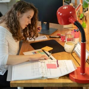 Woman working intently at her desk in a cozy office, surrounded by documents and a bright red lamp.